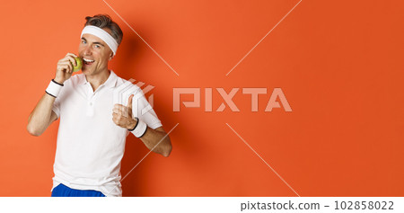 Portrait of healthy and active middle-aged man, eating an apple and showing thumbs-up, standing against orange background 102858022