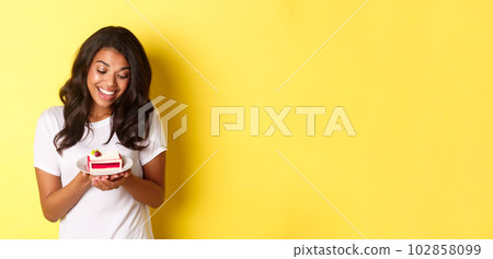 Portrait of attractive african-american woman, looking at delicious piece of cake and smiling, standing over yellow background Portrait of attractive african-american woman, looking at delicious piece of cake and smiling, standing over yellow background 102858099