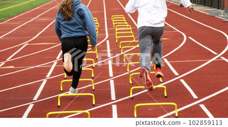 Girl and boy running over small yellow hurdles in lane on a track in the cold 102859113