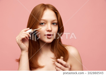 Portrait of young, beautiful, redhead girl with freckled skin applying concealer on face against pink studio background 102860543