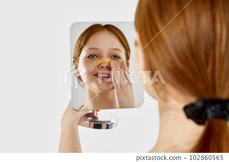 Happy, young, beautiful girl with bandage on her nose after rhinoplasty looking at mirror against white studio background 102860565