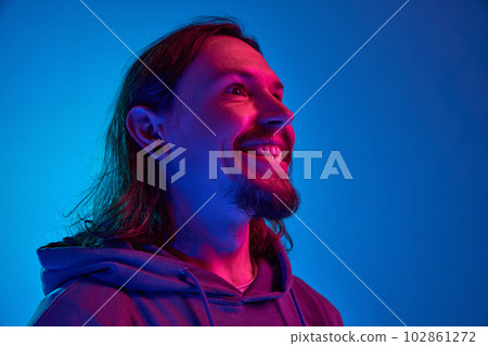 Portrait of man with moustache in casual clothes, hoodie looking at camera, smiling, posing against blue studio background in neon light Portrait of man with moustache in casual clothes, hoodie looking at camera, smiling, posing against blue studio background in neon light 102861272