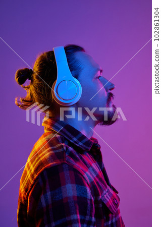 Side view portrait of young man listening to music in headphones with serious face against purple studio background in neon light 102861304
