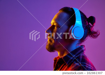Side view portrait of young man listening to music in headphones against purple studio background in neon light Side view portrait of young man listening to music in headphones against purple studio background in neon light 102861307