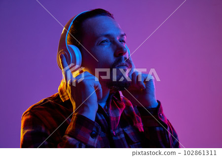 Portrait of young man in checkered shirt listening to music in headphones against purple studio background in neon light Portrait of young man in checkered shirt listening to music in headphones against purple studio background in neon light 102861311