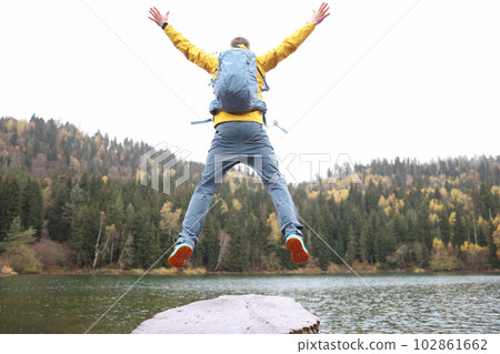 Happy male traveler with backpack jumping near forest lake back view 102861662