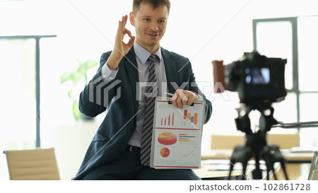 Business coach man showing ok gesture and holding papers with charts in front of camera 102861728