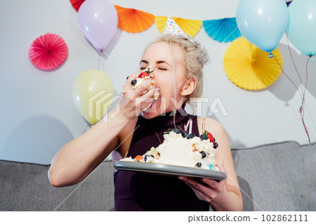 Woman in festive dress and party cap eating birthday cake with her hands, her face stained cream. A woman stuffing her face with cake. Crazy celebrating birthday party at home. Selective focus. Woman in festive dress and party cap eating birthday cake with her hands, her face stained cream. A woman stuffing her face with cake. Crazy celebrating birthday party at home. Selective focus. 102862111