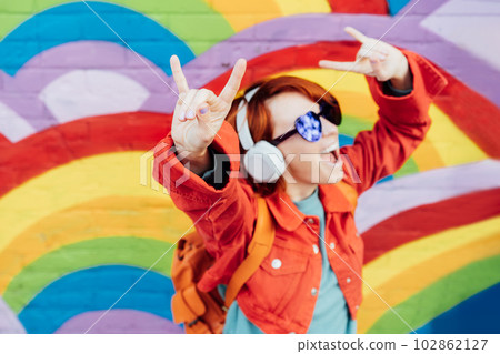 Focus on rock and roll hand gesture of emotional stylish woman wearing wireless headphones, listening to music and singing on rainbow brick wall background. Music festival. hipster lifestyle. Focus on rock and roll hand gesture of emotional stylish woman wearing wireless headphones, listening to music and singing on rainbow brick wall background. Music festival. hipster lifestyle. 102862127