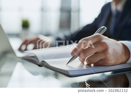 Close up of woman's hands writing in spiral notepad placed on wooden desktop with various items Close up of woman's hands writing in spiral notepad placed on wooden desktop with various items 102862132