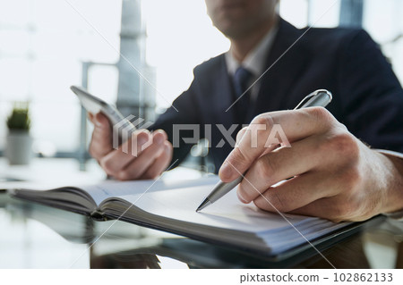 Businessman writes a note on a notepad while looking at a smartphone Businessman writes a note on a notepad while looking at a smartphone 102862133