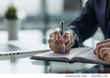 Close up of woman's hands writing in spiral notepad placed on wooden desktop with various items Close up of woman's hands writing in spiral notepad placed on wooden desktop with various items 102862134
