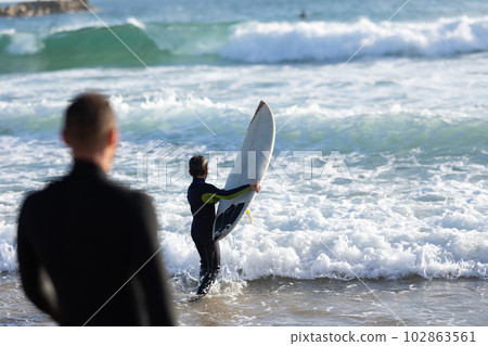 A man watches a little boy going out in the sea holding a surfing board 102863561