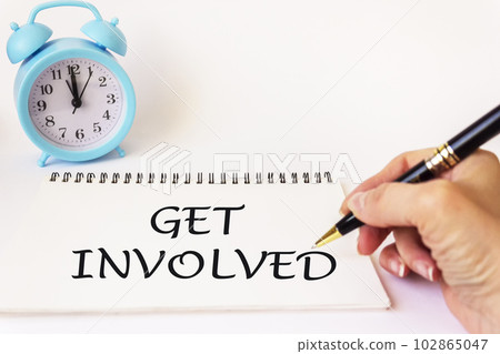 Text Get involved written on a notebook by a female hand, pen to the side on a white background  102865047