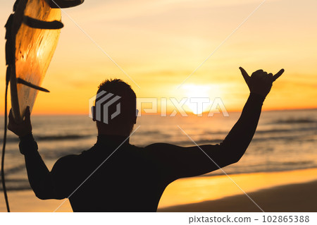 Silhouette of a man holding a surfboard showing shaka at sunset 102865388