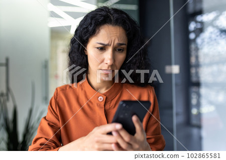 Upset young arab businesswoman sitting in office and looking worriedly at phone. Received bad news by message, checking account, communication problems. Upset young arab businesswoman sitting in office and looking worriedly at phone. Received bad news by message, checking account, communication problems. 102865581
