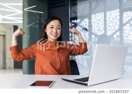 A young Asian business woman working in the office using a laptop, confidently and smilingly looking at the camera and showing a power gesture with her hands. Successfully completed work, project. A young Asian business woman working in the office using a laptop, confidently and smilingly looking at the camera and showing a power gesture with her hands. Successfully completed work, project. 102865587
