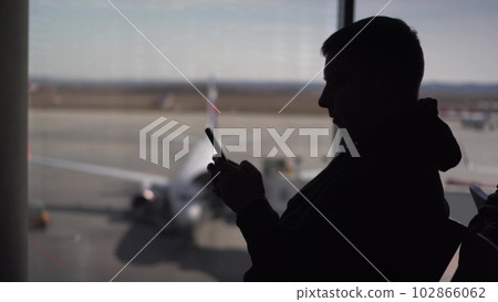 A young man in a sweatshirt is sitting at the airport with a phone in his hands. The man is using a smartphone. Silhouette of a man on the background of the planes outside the window. A young man in a sweatshirt is sitting at the airport with a phone in his hands. The man is using a smartphone. Silhouette of a man on the background of the planes outside the window. 102866062