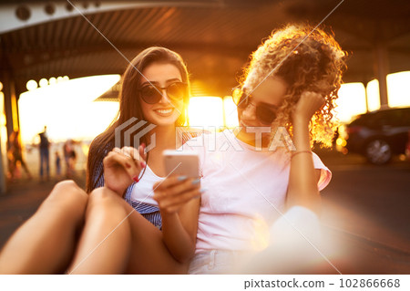 Selfie time! Two beautiful girls having fun on shopping trolleys. Multiethnic young people racing on shopping cart. Selfie time! Two beautiful girls having fun on shopping trolleys. Multiethnic young people racing on shopping cart. 102866668