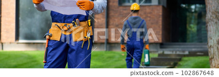 Cropped shot of male builder in blue overalls standing outdoors with open blueprint while working on cottage construction Cropped shot of male builder in blue overalls standing outdoors with open blueprint while working on cottage construction 102867648