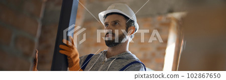Focused young male builder wearing hard hat holding metal stud for drywall while working at construction site 102867650
