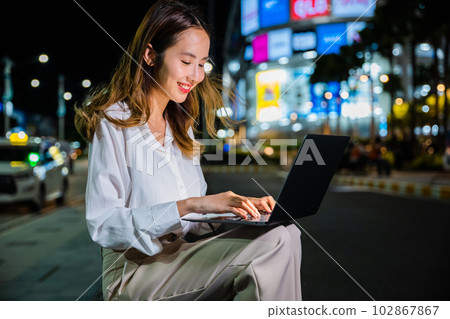 Amidst the busy city nightlife, a woman is hard at work on her laptop computer, utilizing the connectivity and convenience of modern technology to keep up with her online business and communication. 102867867