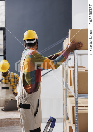 African american storehouse employee taking cardboard box for order packing from shelf. Warehouse worker wearing overall and yellow helmet standing on ladder with carton in hands African american storehouse employee taking cardboard box for order packing from shelf. Warehouse worker wearing overall and yellow helmet standing on ladder with carton in hands 102868327