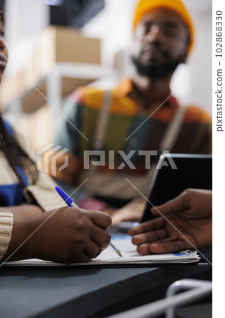 African american woman warehouse worker hand writing in clipboard at reception desk. Storehouse employees checking order list and managing documentation at post office checkout close up African american woman warehouse worker hand writing in clipboard at reception desk. Storehouse employees checking order list and managing documentation at post office checkout close up 102868330