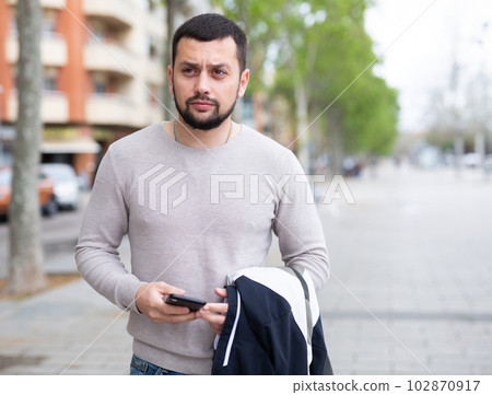 Focused guy with jacket and phone in hands on city street 102870917
