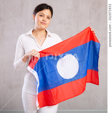 Middle-aged positive woman in casual clothes holds unfurled flag of Laos in hands raised above head Middle-aged positive woman in casual clothes holds unfurled flag of Laos in hands raised above head 102871652