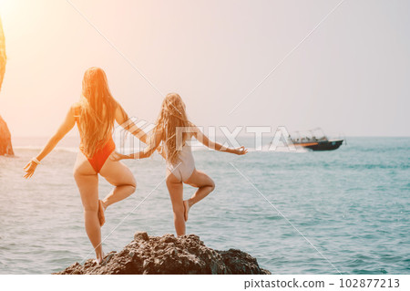 Woman and her daughter practicing balancing yoga pose on one leg up together on rock in the sea. Silhouette mother and daughter doing yoga at beach 102877213