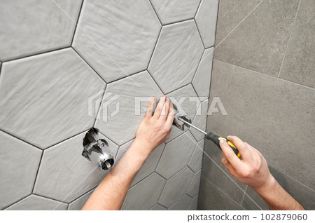 Close up of plumber using screwdriver while installing shower system components into wall with ceramic tile. Man screwing metal bolt with plumbing repair tool while working on bathroom renovation. Close up of plumber using screwdriver while installing shower system components into wall with ceramic tile. Man screwing metal bolt with plumbing repair tool while working on bathroom renovation. 102879060
