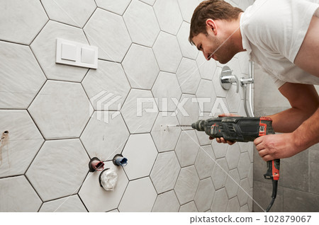 Side view of male worker drilling wall with cordless power drill in apartment under renovation. Man using electric drilling machine while working on bathroom renovation and installing shower system. 102879067