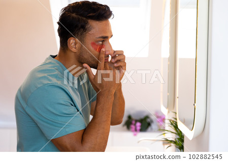 Biracial man looking in mirror and applying under eye mask in bathroom Biracial man looking in mirror and applying under eye mask in bathroom 102882545
