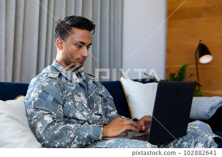 Biracial male soldier wearing military uniform, sitting on sofa using laptop at home Biracial male soldier wearing military uniform, sitting on sofa using laptop at home 102882641