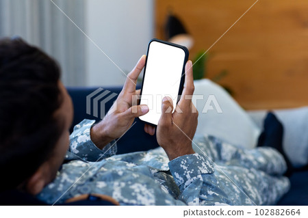 Biracial male soldier wearing uniform, lying on sofa using smartphone, with copy space on screen 102882664