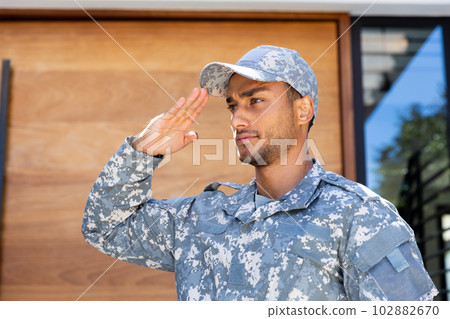 Biracial male soldier wearing uniform and cap, saluting outside house 102882670