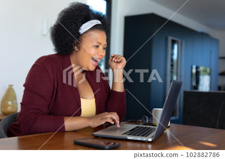 Happy plus size african american woman sitting at table, making video call using laptop Happy plus size african american woman sitting at table, making video call using laptop 102882786