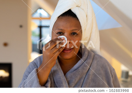 Happy plus size african american woman wearing robe, cleansing face with cotton pad in bathroom Happy plus size african american woman wearing robe, cleansing face with cotton pad in bathroom 102883998