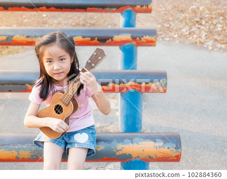Little asian child girl play the ukulele, in the garden on the Steel pipe, practice to play Little asian child girl play the ukulele, in the garden on the Steel pipe, practice to play 102884360