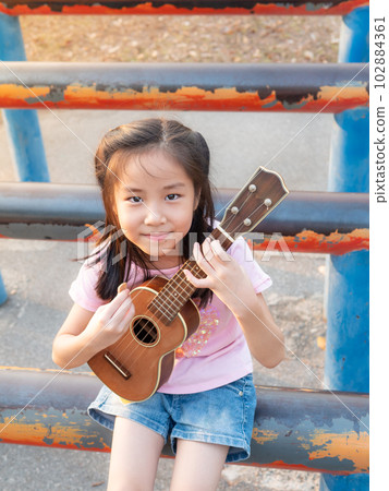Little asian child girl play the ukulele, in the garden on the Steel pipe, practice to play Little asian child girl play the ukulele, in the garden on the Steel pipe, practice to play 102884361