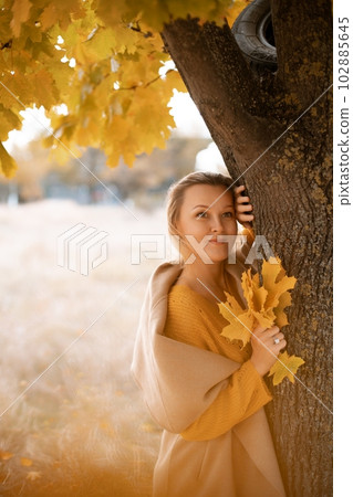 The blonde stands near the autumn tree. Thoughtful woman looks ahead, dressed in a yellow dress. Autumn content 102885645