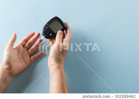 Hands of caucasian woman taking blood sugar reading with glucometer, on blue background, copy space 102888462