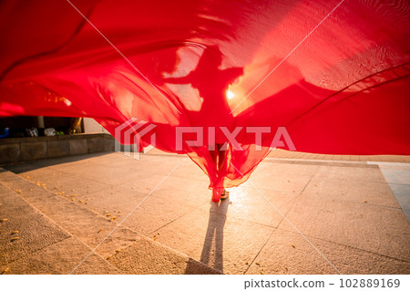 Sunrise red dress. A woman in a long red dress against the backdrop of sunrise, bright golden light of the sun's rays. The concept of femininity, harmony. 102889169