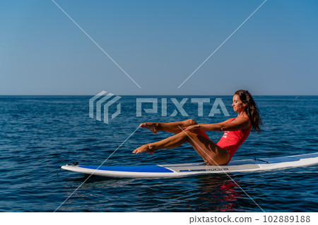 Sporty girl on a surfboard in the sea on a sunny summer day. In a red swimsuit, she sits in the splits on the sap. Summer entertainment on Stortom by the sea 102889188