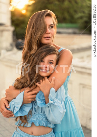 Mother daughter sunset. in blue dresses with flowing long hair against the backdrop of sunset. The woman hugs and presses the girl to her. 102889216