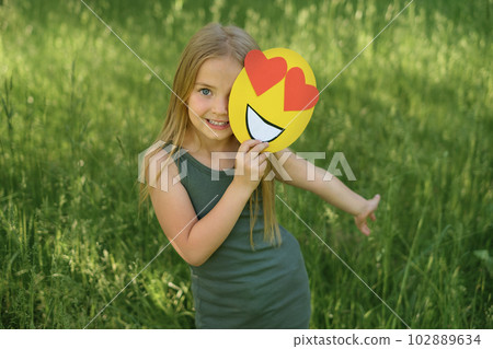 A beautiful darling sweet  blonde girl holds in her hand a paper face of a loving smile with hearts instead of eyes. 102889634