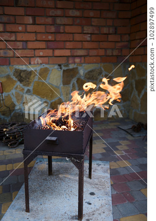 Still life with branches burning in flaming fire in barbecue grill. Preparation of food in the open air in the backyard against a red brick wall background. The season of summer picnic and BBQ party 102897499