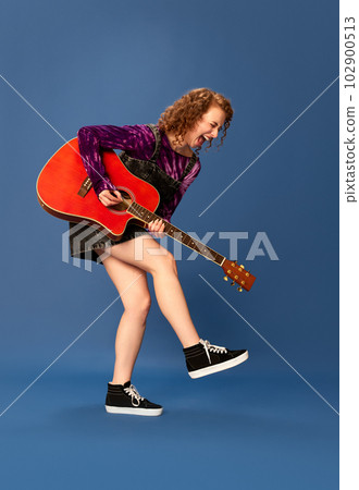 Artistic musician. Burst of energy. Portrait of young emotional girl playing guitar, having fun against blue studio background 102900513
