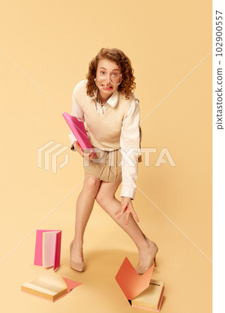 Portrait of young girl with funny face in image of student or trainee, in glasses, with many books against studio background Portrait of young girl with funny face in image of student or trainee, in glasses, with many books against studio background 102900557